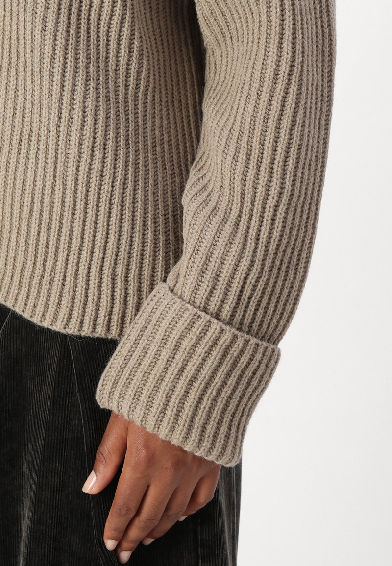 Beige gerippter Strickpullover mit breiten Bündchen. Die Textur ist dick und zeigt vertikale Linien sowie ein gemütliches Erscheinungsbild. Kombiniert mit dunklem Cord.