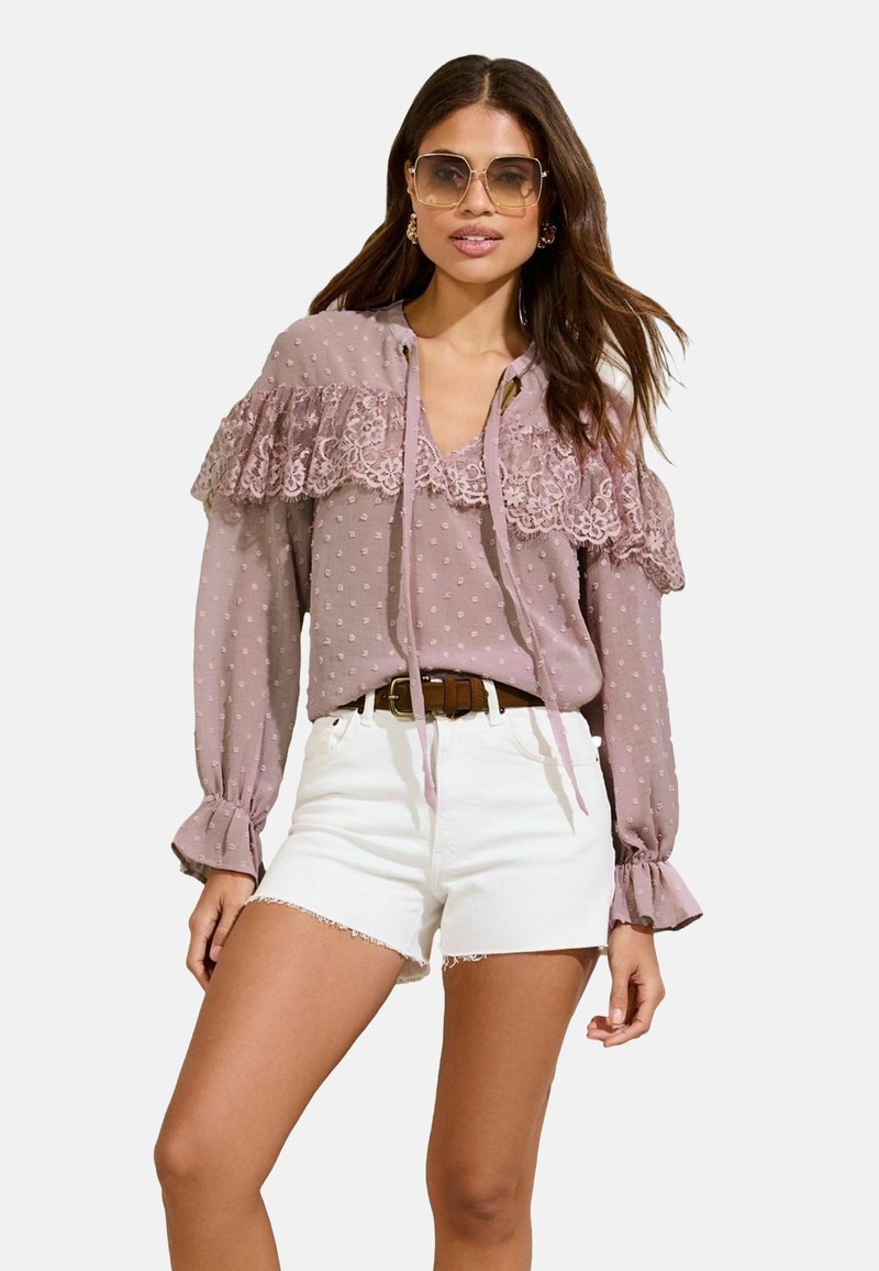Light mauve blouse with polka dots and lace details, featuring a tie neckline and ruffled sleeves, paired with white denim shorts and a brown belt.