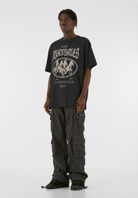 Schwarzes Grafik-T-Shirt mit beigen Text und Globusdesign, kombiniert mit dunklen Cargo-Hosen mit mehreren Taschen und entspannter Passform.