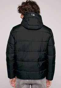 Chaqueta acolchada negra con capucha, que presenta costuras horizontales, un brillo sutil y detalle del logo en la parte superior de la espalda. Tejido texturizado con un acabado suave.