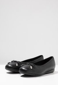 Bailarinas de cuero negro con punta redondeada, con un detalle de lazo y pequeños acentos plateados. Textura suave con una suela flexible.