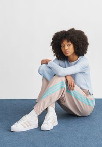 Mulher com cabelo encaracolado sentada de pernas cruzadas num tapete azul, vestindo uma camisola azul clara, calças bege com riscas azuis, meias brancas e ténis brancos.