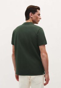 Camisa polo verde oscuro, de manga corta, diseño liso, tejido texturizado, con aberturas laterales en el dobladillo, llevada con pantalones de color claro, vista desde la parte de atrás.