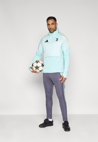 Lyseblå fodbold-hættetrøje med pink accenter og sort logo, kombineret med grå slim-fit bukser med blå sidestriber. Holder en mønstret fodbold.