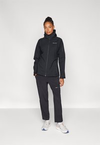 Black waterproof jacket with a zip front, hood, and side pockets, paired with black pants featuring zippered pockets and light sneakers.