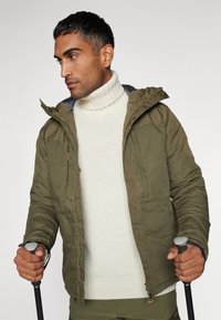 Olijfgroene gewatteerde jas met capuchon, gecombineerd met een crèmekleurige dikke gebreide coltrui. De jas heeft zijzakken en een ritssluiting.