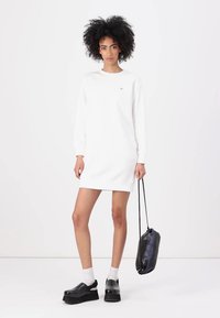 Vestito bianco a felpa con maniche lunghe e scollo rotondo. Abbinato a zoccoli neri, calze bianche e una borsa nera con cordoncino.