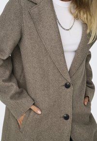 Manteau en feutre marron avec col à revers, trois boutons noirs, poches latérales et texture lisse. Porté par-dessus un haut blanc avec une chaîne argentée.