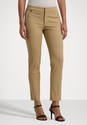 Femme portant un pantalon beige slim et des sandales noires à talons hauts, debout sur un fond blanc uni.