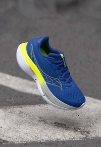 Baskets de course en bleu avec une tige perforée, des accents jaunes et une semelle blanche. Présente un design fluide et un marquage visible.