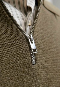 Pruun tõmblukuga kampsun, millel on tekstuuriga kudumismuster, esitatud hõbedase tõmbluku ja ribaga särgi nähtavusega allpool. Tõmblukk on bränditud.