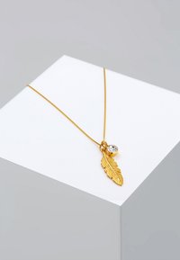 Gouden ketting met een bladvormig hangertje met gedetailleerde textuur en een kleine heldere steen, gelegen op een geometrische display.