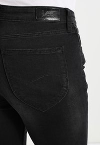 Vaqueros de mezclilla negra con un bolsillo trasero que presenta un detalle de costura curvada y un parche de logo de piel en la cinturilla. Textura suave.