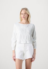 Blusa bianca a maniche lunghe con pieghe testurizzate e dettagli ricamati, abbinata a pantaloncini di jeans bianchi con orli sfilacciati.