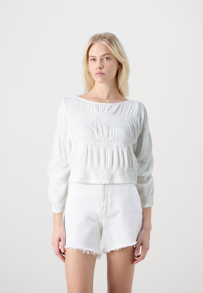 Weiße Bluse mit langen Ärmeln, strukturierten Falten und bestickten Akzenten, kombiniert mit weißen Denim-Shorts mit fransigen Kanten.