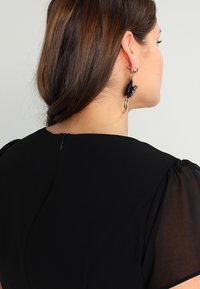 Svart plagg med ett slätt tyg och genomskinliga puffärmar. Har en diskret dragkedja på ryggen. Långa örhängen med dekorativ detalj.