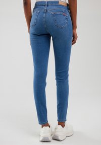 Hátsó nézet egy személyről, aki vékony kék farmert és fehér, masszív sportcipőt visel, egyszínű háttér előtt.