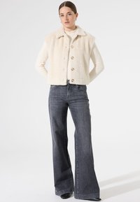 Crèmekleurige, pluche, mouwloze jas met een kraag en vijf knopen. In combinatie met hoog gewaiste, flared, grijze jeans. Eenvoudig ontwerp, zachte textuur.