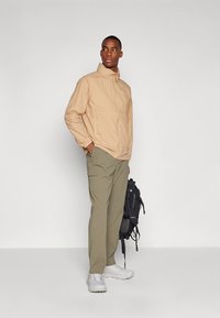 Chaqueta ligera beige con cuello alto, combinada con pantalones cargo verde oliva. El modelo sostiene una mochila negra y lleva zapatillas blancas.