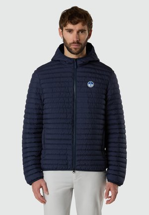 Hombre con chaqueta acolchada azul marino con capucha y cremallera cerrada, con parche de logo de brújula en el pecho izquierdo y pantalones de color claro.