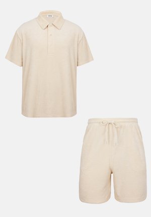 Beiges kurzärmeliges Polohemd mit Knopfleiste, kombiniert mit beigen Shorts mit Kordelzug vor einfarbigem Hintergrund.