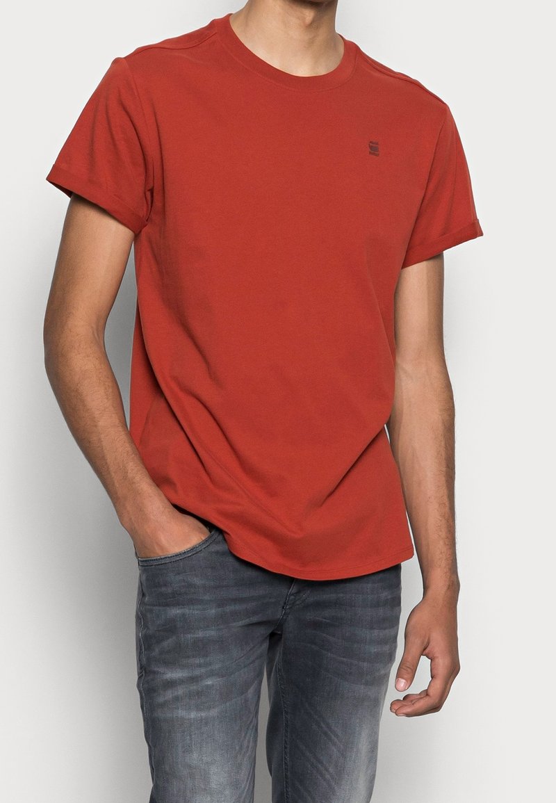 Personne portant un t-shirt rouge uni à manches courtes avec un petit logo sur la poitrine, assorti à un jean bleu foncé délavé.