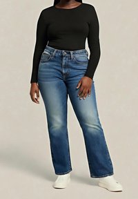 Jeans évasés bleus avec effet délavé clair, design à cinq poches, accessoires en métal et taille haute. Associés à un haut noir cintré à manches longues.