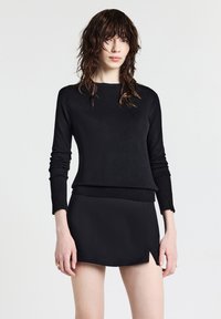 Donna con capelli scuri e bagnati, che indossa un maglione nero a maniche lunghe e una minigonna nera con una piccola fessura frontale.