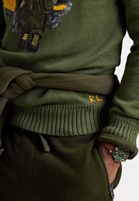 Maglione verde lavorato a maglia con un design ricamato che presenta una figura e dettagli gialli. Polsini e orlo a costine, abbinato a joggers color oliva.