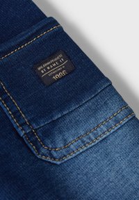 Donkerblauwe denimstof met een gestructureerd oppervlak. Voorzien van een label met de tekst "by NAME IT" en contrasterende gouden stiksels langs de randen.