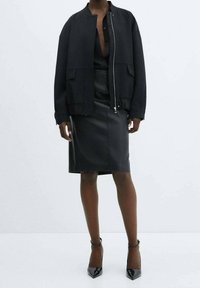 Veste aviateur noire avec fermeture éclair, deux poches avant ; associée à une jupe en cuir noir moulante et des talons en cuir verni.