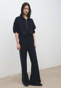 Temno modra jopa z zadrgo in zavihanimi manšetami, združena z ujemajočimi širokimi hlačami. Značilne so bele stranske črte in gladka tekstura.