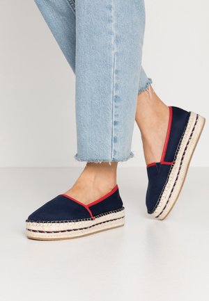 Fødder iført marineblå espadrille platformssko med rød kant og lyseblå korte jeans på en ensfarvet hvid gulv- og baggrund.