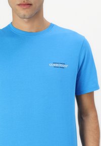 Trumpas mėlynas T-shirt pagamintas iš medvilnės, su apvalia kaklo iškirpte ir mažu prekės ženklu krūtinėje balta ir juoda spalvomis.