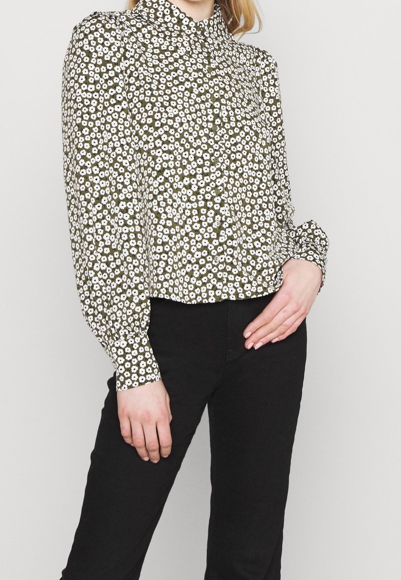 Femme portant un chemisier olive à manches longues avec un motif floral blanc et un pantalon noir, la main reposant sur la hanche devant un fond uni.