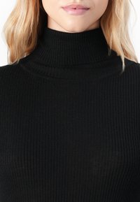 Haut à col roulé noir en maille côtelée avec un design ajusté, mettant en avant un col haut et un tissu texturé. Une vue en gros plan souligne l'élasticité du matériau.