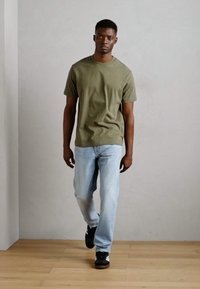 Olive green cotton t-shirt paired with light blue straight-leg jeans. Model wears black sneakers, standing against a neutral backdrop.