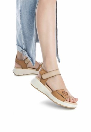 Sandalias de cuero marrón con una correa de velcro, una suela blanca acolchada y un logo destacado en la zona del dedo, combinadas con denim desgastado.