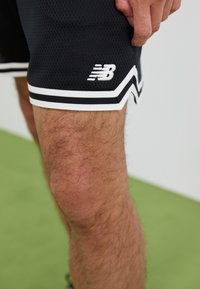 Shorts de sport noirs avec une texture en maille, accents blancs et un logo New Balance sur le côté inférieur. Présente un design d'ourlet contrastant.
