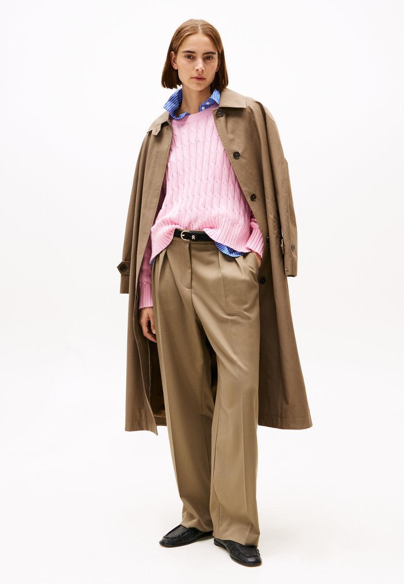 Beige trenchcoat, lys rosa kabelstrikket genser, blå skjorte under, brune brede bukser, svarte sko. Glatte teksturer, lagdelt stil.