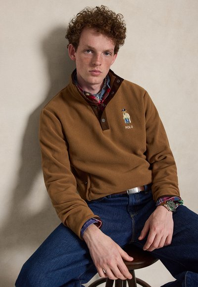 Νέος άνδρας με σγουρά μαλλιά φοράει καφέ fleece Polo, καρό πουκάμισο, μπλε τζιν και ρολόι χειρός, καθισμένος σε σκαμπό μπροστά από έναν απλό τοίχο.