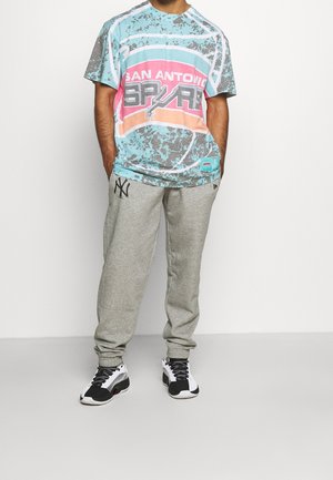 Personne portant un T-shirt multicolore des San Antonio Spurs, un pantalon de survêtement gris avec un logo noir, et des baskets noir et blanc, se tenant contre un fond blanc.