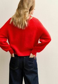 Röd ulltröja med oversized passform, ribbade muddar och nederkant. Kombinerad med mörkblå vida jeans. Bakifrån visas den avslappnade stilen.