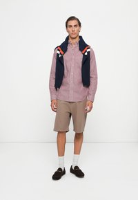 Rood-witte gestreepte lange mouwen shirt, beige patroon shorts, marineblauwe trui over de schouders, bruine instappers en witte crew-sokken.