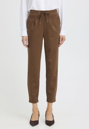 Nő barna, szűk szárú, húzózsinóros derekú nadrágban és fehér hosszú ujjú ingben, sötét, hegyes orrú cipővel, fehér háttér előtt állva.