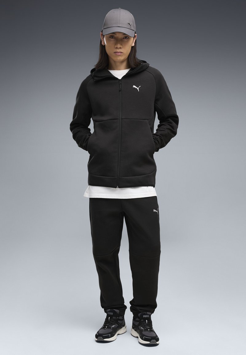 Jeune personne portant un sweat à capuche noir Puma, un pantalon noir, une casquette grise et des baskets noires Puma, debout les mains dans les poches sur fond gris.