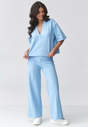Kobieta w luźnej, jasnoniebieskiej bluzce z dekoltem w serek i pasujących szerokich spodniach, pozująca i uśmiechająca się na jednolitym tle.