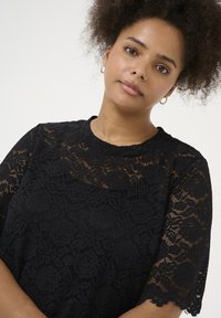 Black lace top featuring a floral pattern, round neckline, and short sleeves with scalloped edges. Lightweight texture and intricate detailing.