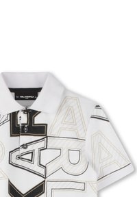 Fehér póló, fekete és arany geometrikus betűzéssel, 3 gombos záródással és texturált anyaggal. Márkájának címkéje látható a gallér belsejében.