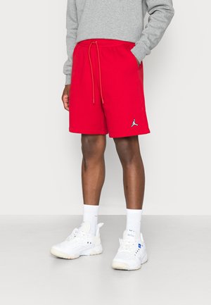 Osoba nosząca jaskrawoczerwone sportowe szorty ze sznurkiem i małym logo, w połączeniu z białymi skarpetkami i białymi trampkami.